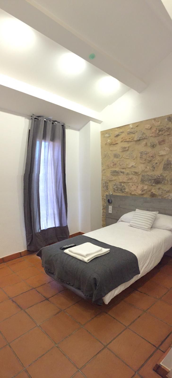Une chambre à coucher avec un lit, un mur en pierre, une fenêtre avec des rideaux et un sol carrelé.