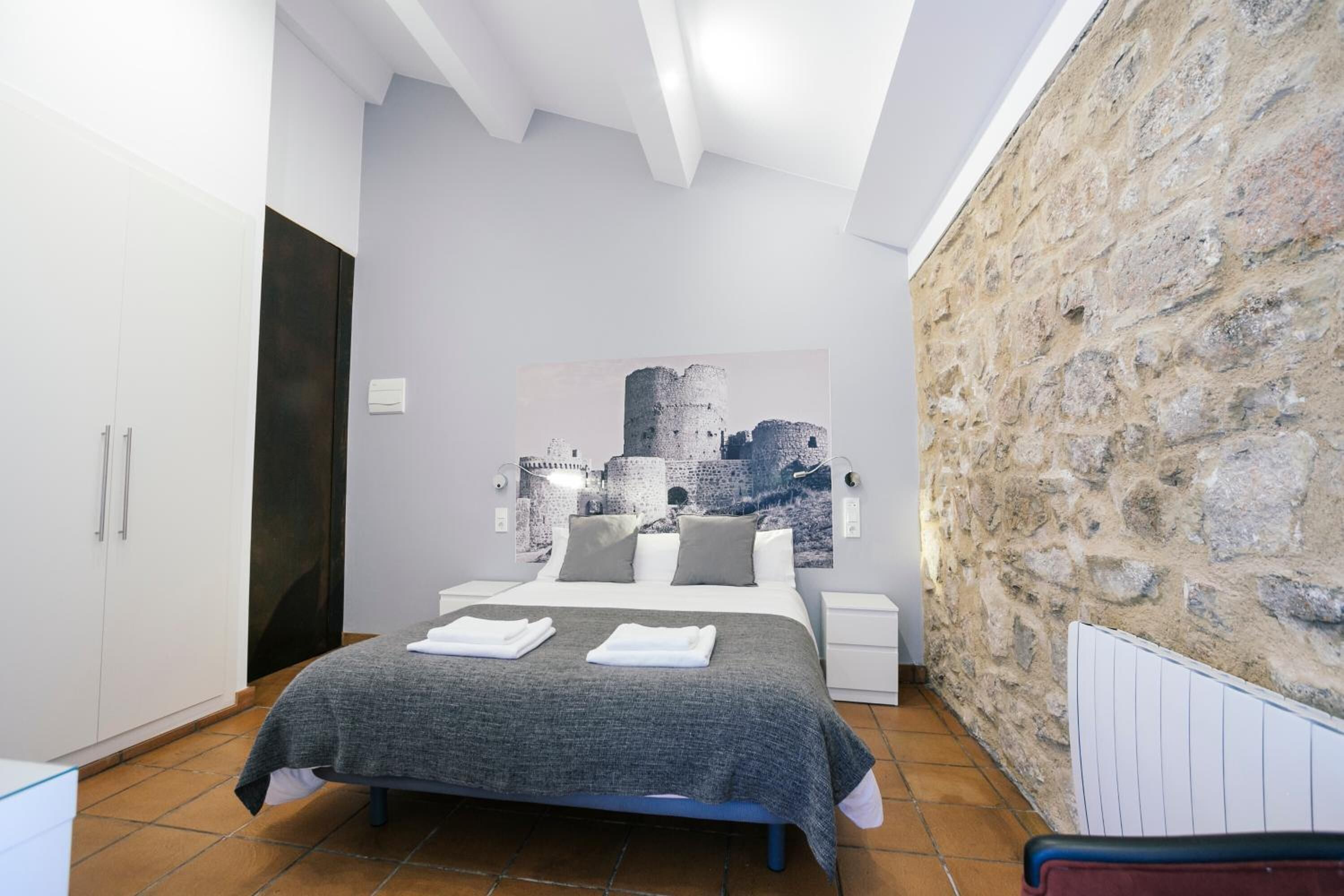 A bedroom with a bed, white wardrobe, stone wall, and a painting of a castle.