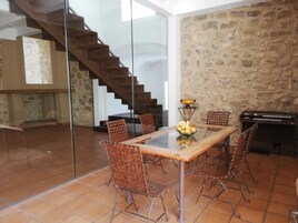 Un comedor con mesa de vidrio, sillas de mimbre, pared de piedra, piano y una escalera.
