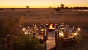 A circular wooden deck with a fire pit and seating, surrounded by a rope fence, overlooking a savanna landscape at sunset.