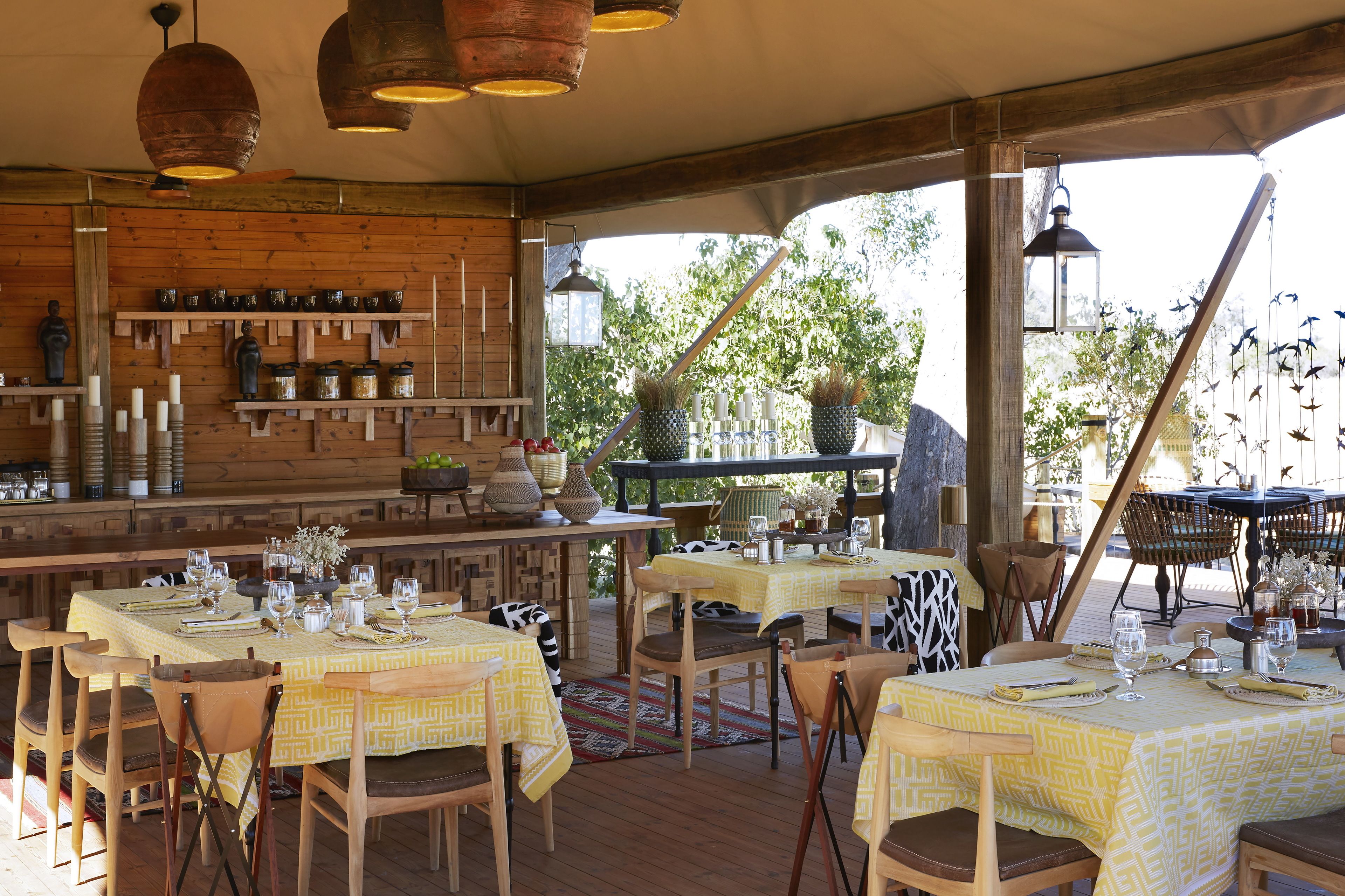 Un espace extérieur aménagé pour les repas, avec des tables et des chaises en bois, protégé par un auvent. On y trouve des lanternes suspendues et divers objets décoratifs accrochés aux murs.