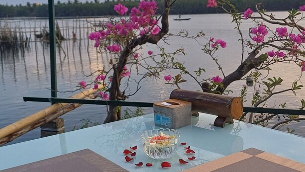 Un tavolo all'aperto con un portatovaglioli di carta e un centrotavola decorativo, sullo sfondo un specchio d'acqua e un albero con fiori rosa.