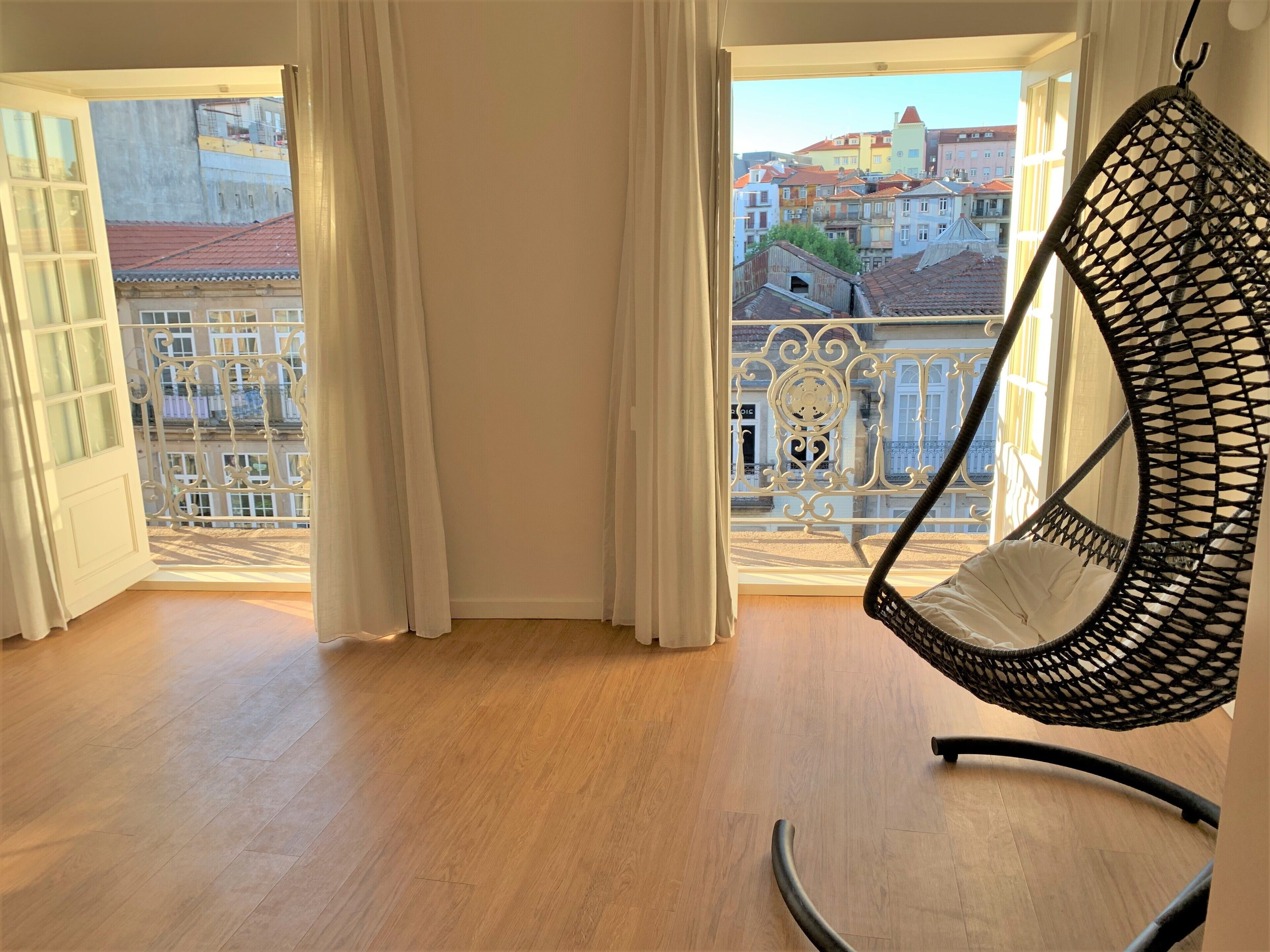 Une pièce dotée d’un plancher en bois, d’un fauteuil suspendu et offrant une vue sur des bâtiments aux couleurs vives, visibles depuis un balcon agrémenté de rideaux blancs.