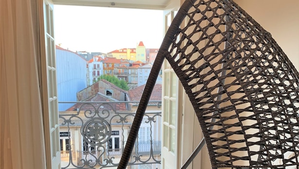 Una silla colgante de mimbre con cojín, ubicada en una habitación con vistas a un edificio a través de un ventanal abierto.