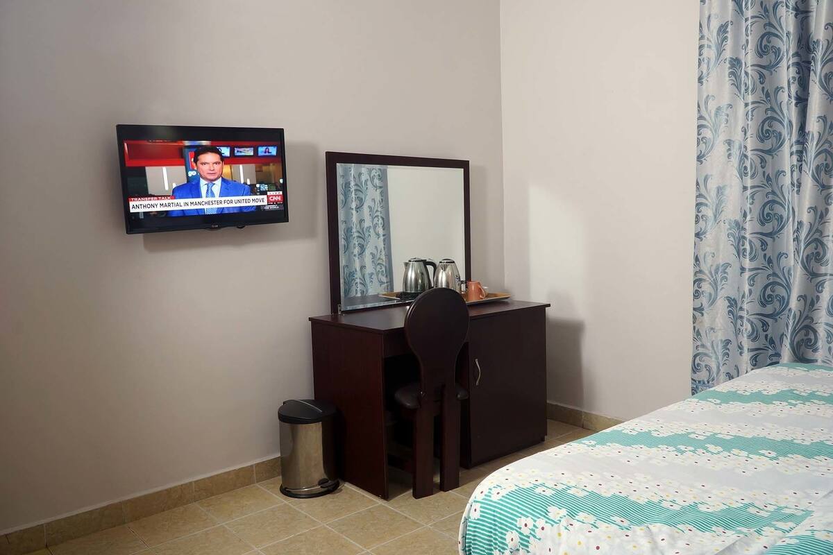 A hotel room with a TV on the wall, a desk with a mirror, a trash bin, and a bed with a patterned bedspread.