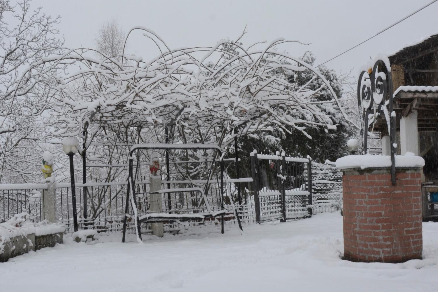 Área externa coberta de neve, com grade de metal, um banco e um pilar de tijolos.