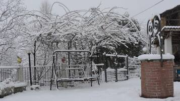 Área externa coberta de neve, com grade de metal, um banco e um pilar de tijolos.