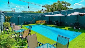 Uma área de piscina com piscina azul cristalina, rodeada por grama sintética verde, e mobiliada com mesas e cadeiras.