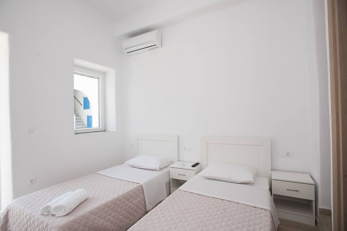 A twin bedroom with two beds, a window, a nightstand, and a wall-mounted air conditioner.