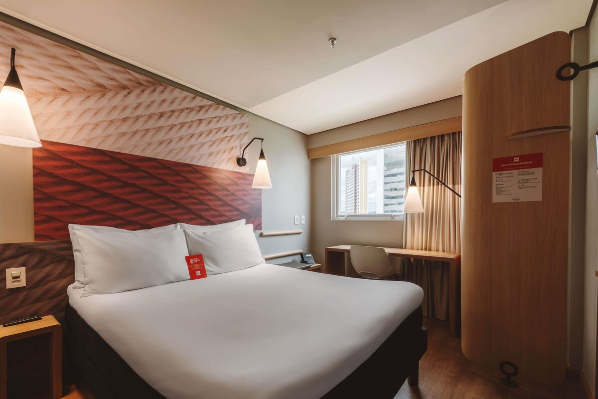 Una habitación de hotel con cama, mesita de noche, lámpara, escritorio, silla y ventana con cortinas.