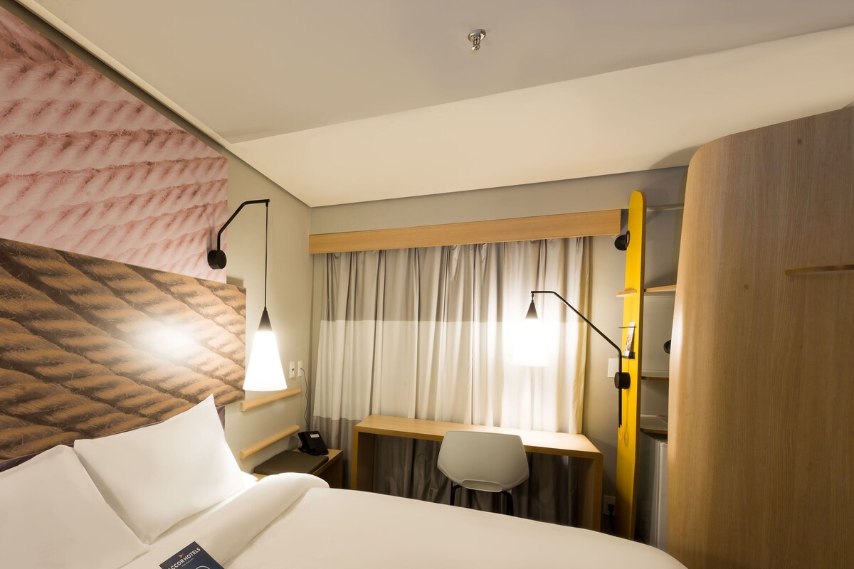 A hotel room with a bed, a desk, a chair, a lamp, and a curtain.