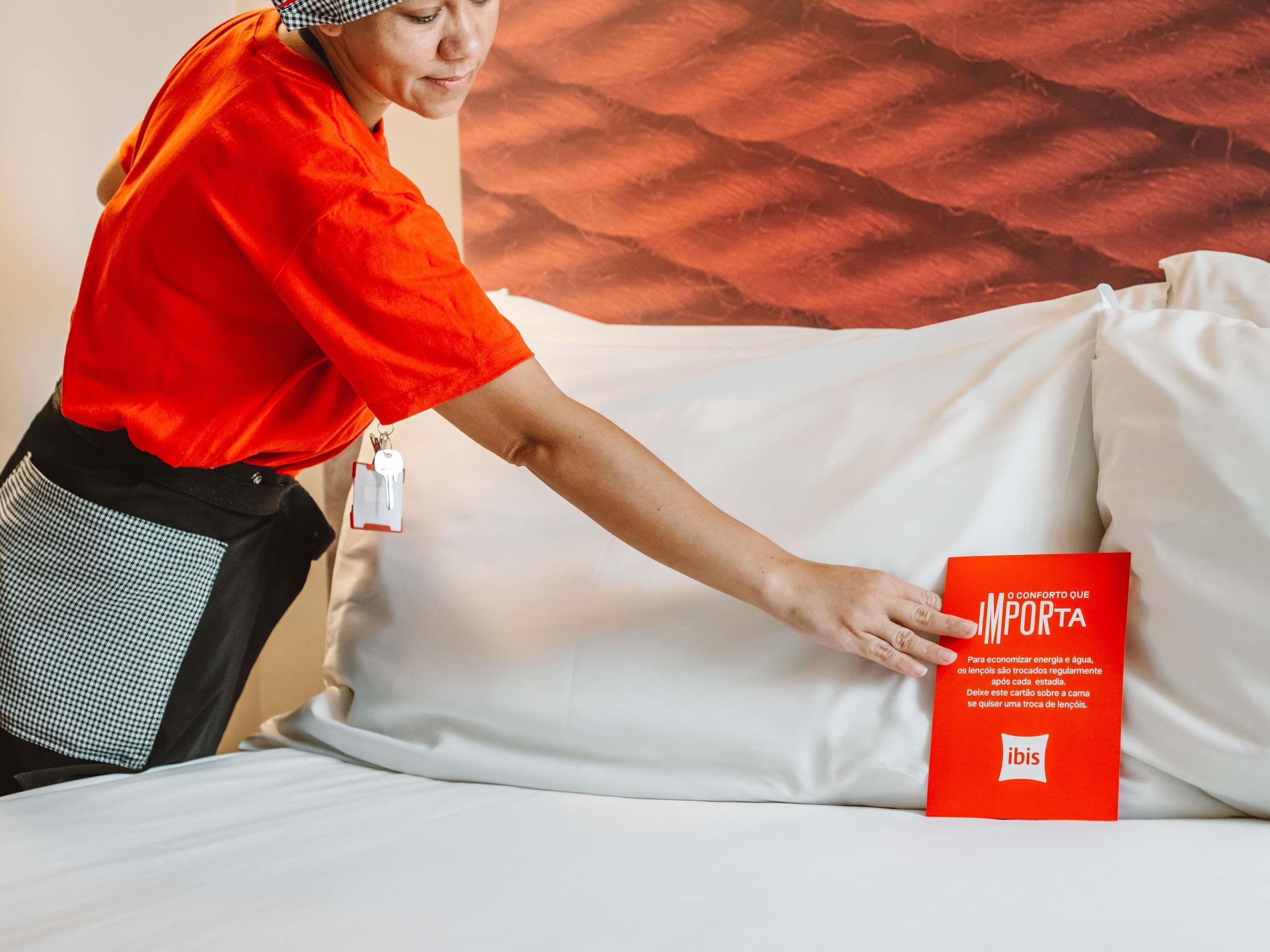 A person in a red uniform adjusting a white pillow on a bed with a red informational card.