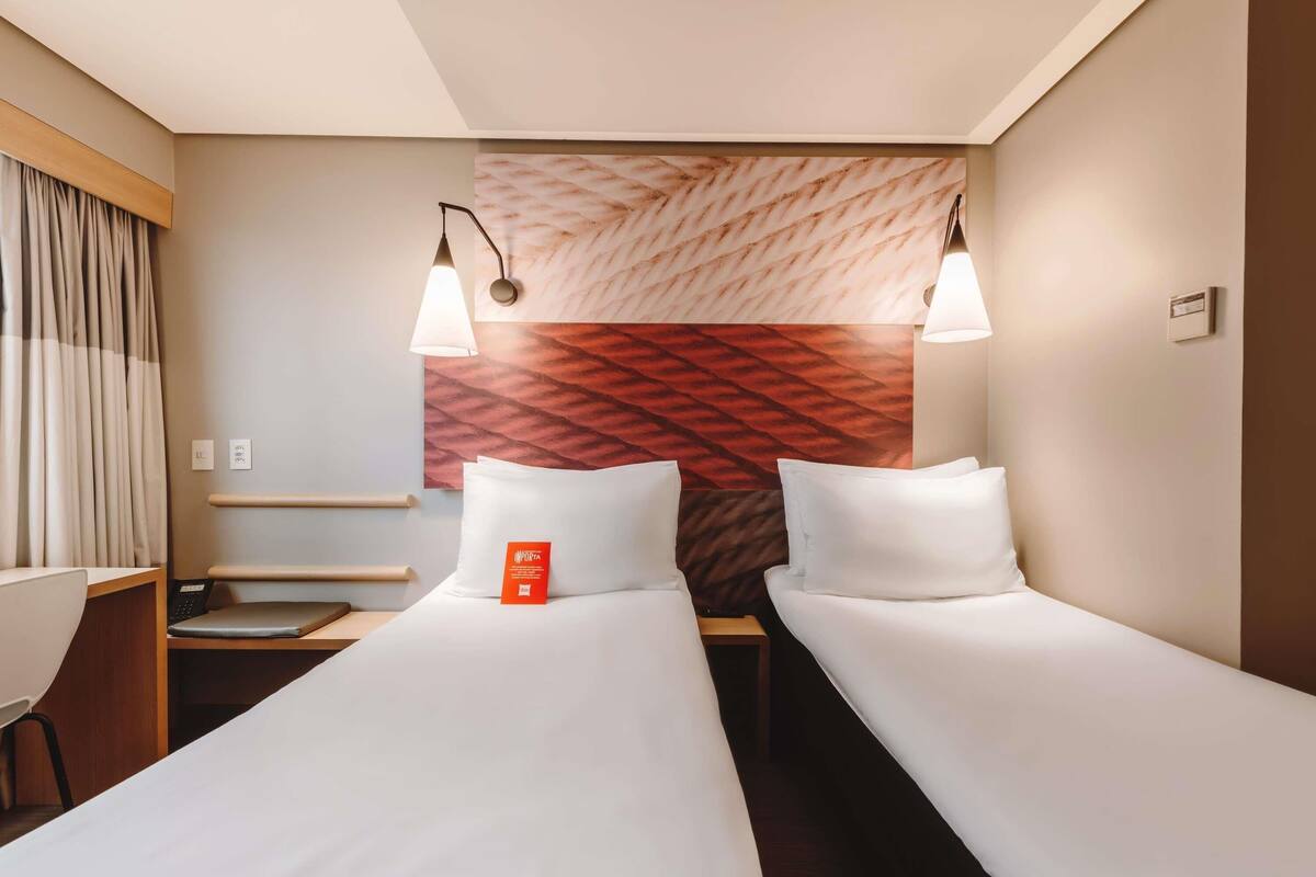 Ein Hotelzimmer mit zwei Einzelbetten, einem Schreibtisch, einem Stuhl und einer Wandlampe.