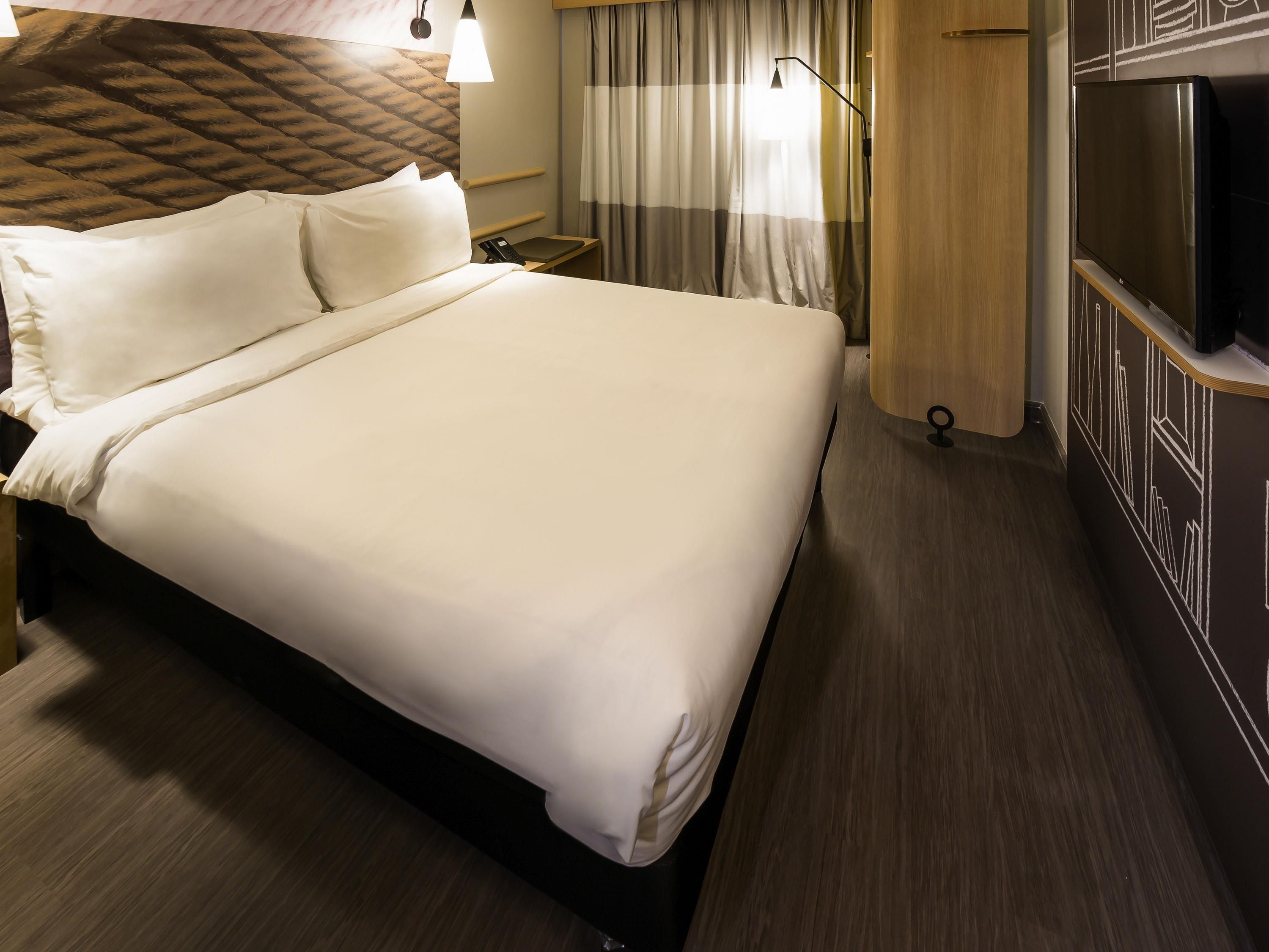 A hotel room with a large bed, a bedside table with a lamp, a TV, and a wooden headboard.