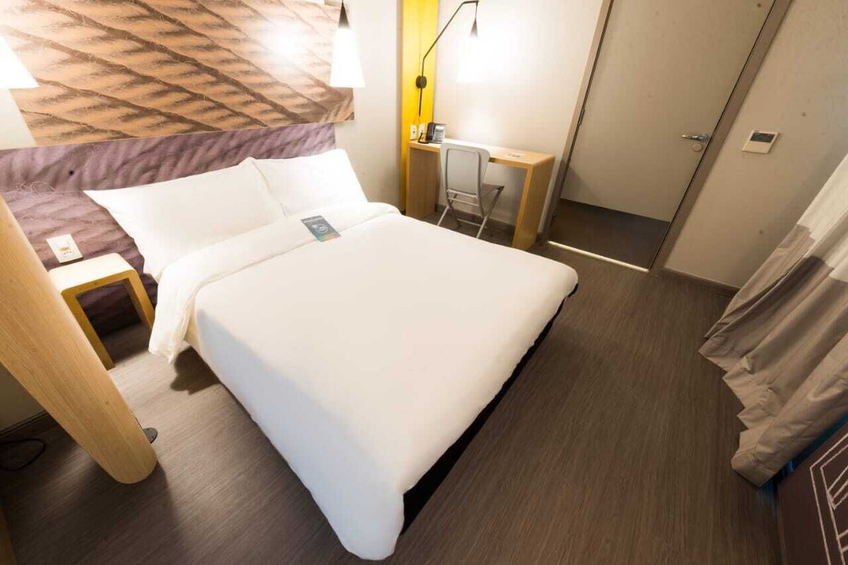 A hotel room with a large bed, a desk, a chair, and a wardrobe.