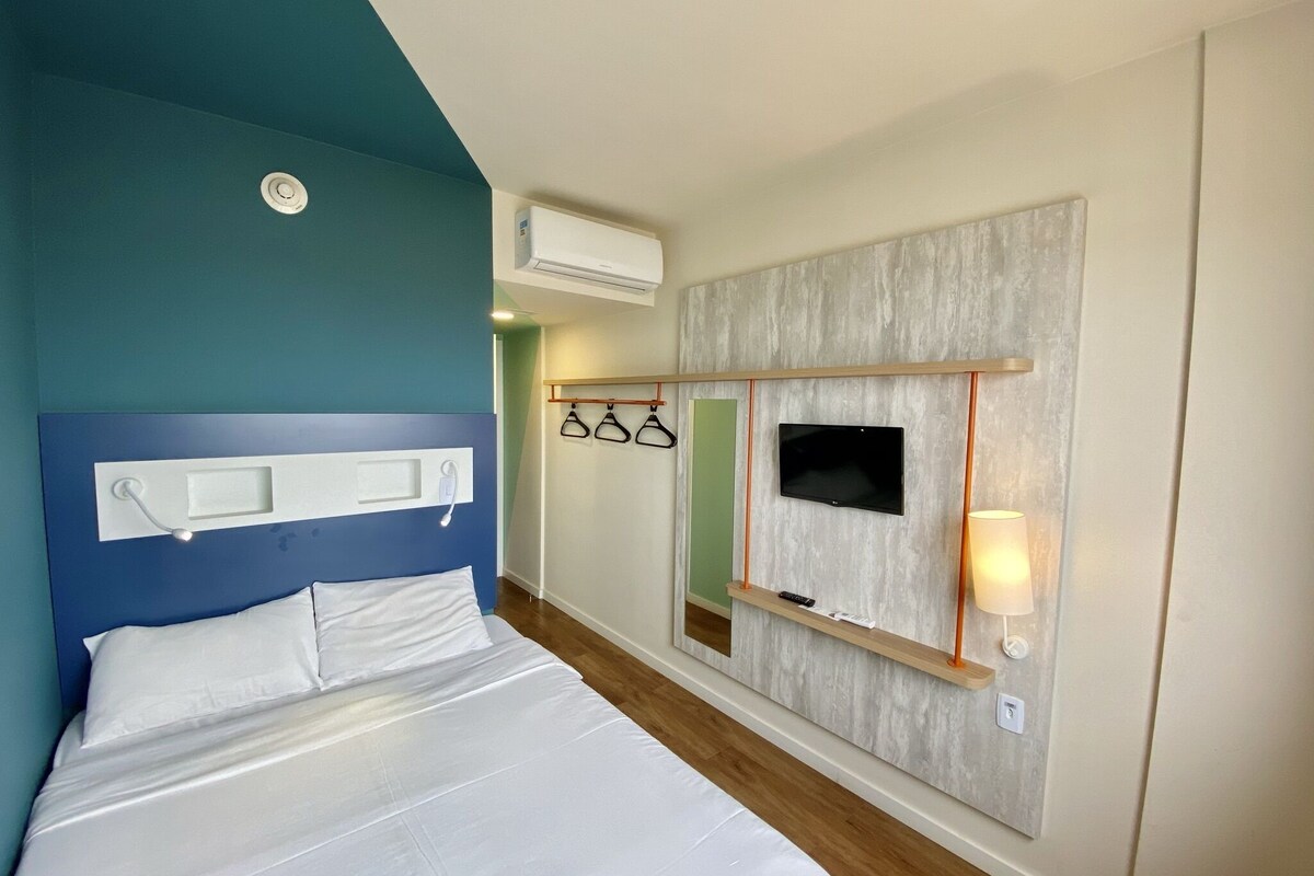 A hotel room with a bed, a mounted TV, a wall-mounted air conditioner, and a mirror with a shelf.