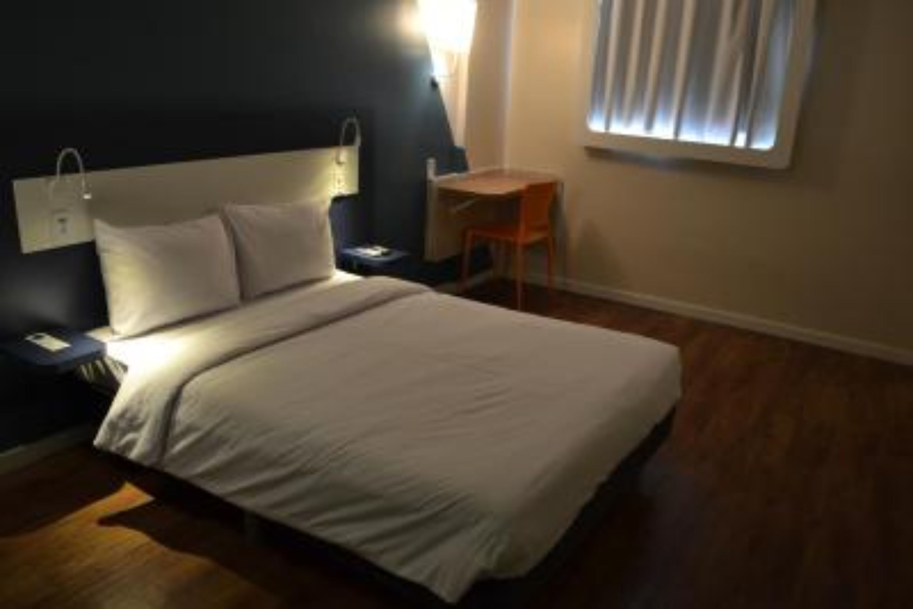 Quarto de hotel com cama, mesa de cabeceira, abajur e uma pequena mesa com uma cadeira.