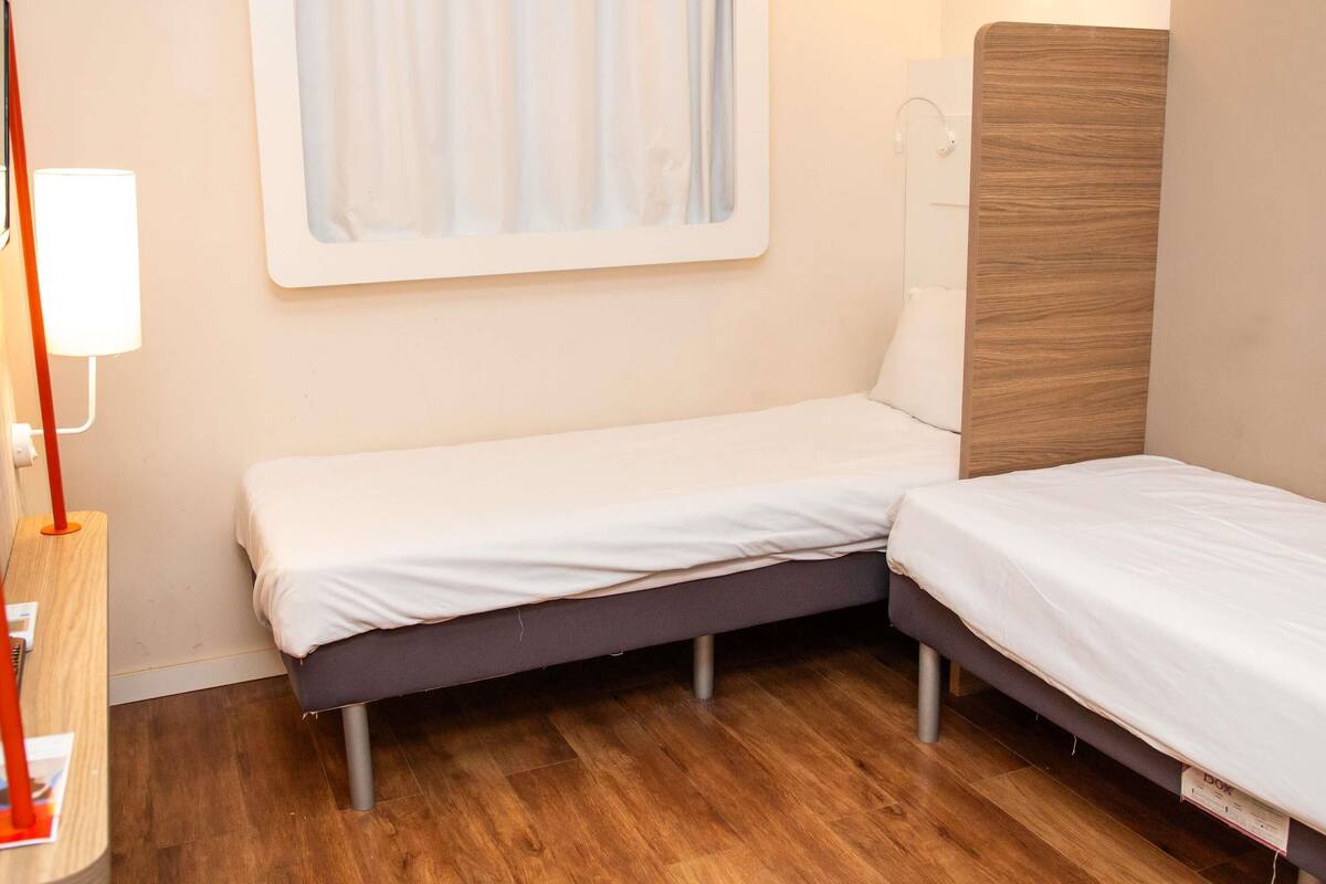 A small, neatly arranged bedroom with a single bed, a bedside table, a lamp, and a window with curtains.