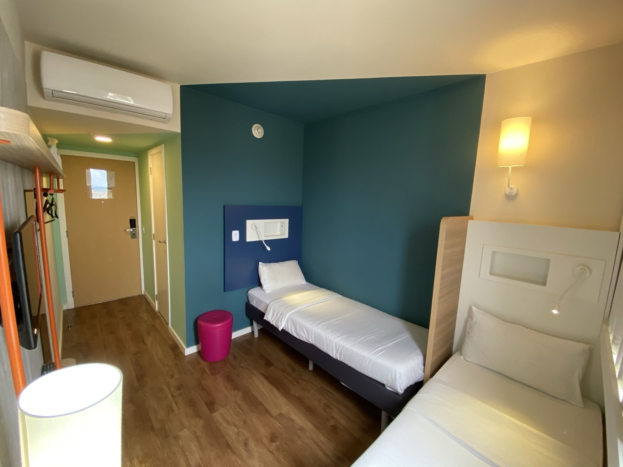 Habitación pequeña y acogedora con dos camas individuales, suelo de madera, pared azul como detalle y un pequeño cubo de basura, además de un teléfono instalado en la pared.