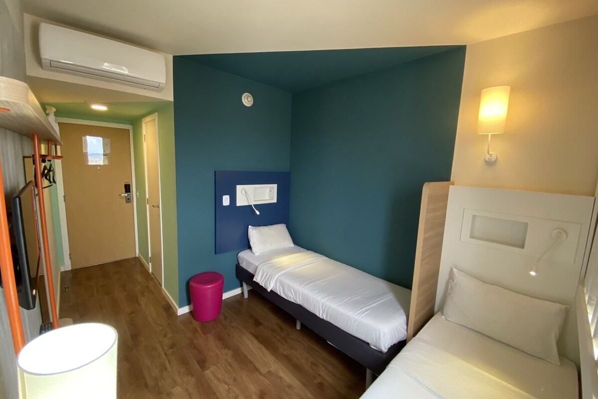 A small, compact room with two single beds, a wooden floor, a blue accent wall, a small trash bin, and a wall-mounted phone.