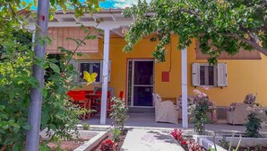Un bâtiment jaune doté d’un patio couvert, d’un espace extérieur aménagé pour s’asseoir et d’un jardin où poussent diverses plantes et fleurs.