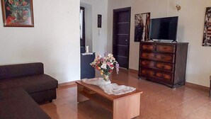Un salon comprenant un canapé, une table basse en bois ornée d’un vase de fleurs, un téléviseur posé sur un meuble et une commode.