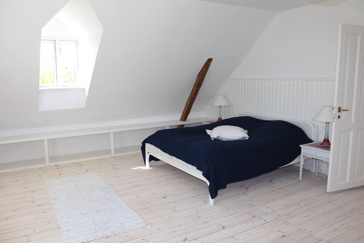 Ein Schlafzimmer mit Bett, Nachttisch, Lampe, Fenster und schräg verlaufender Decke.