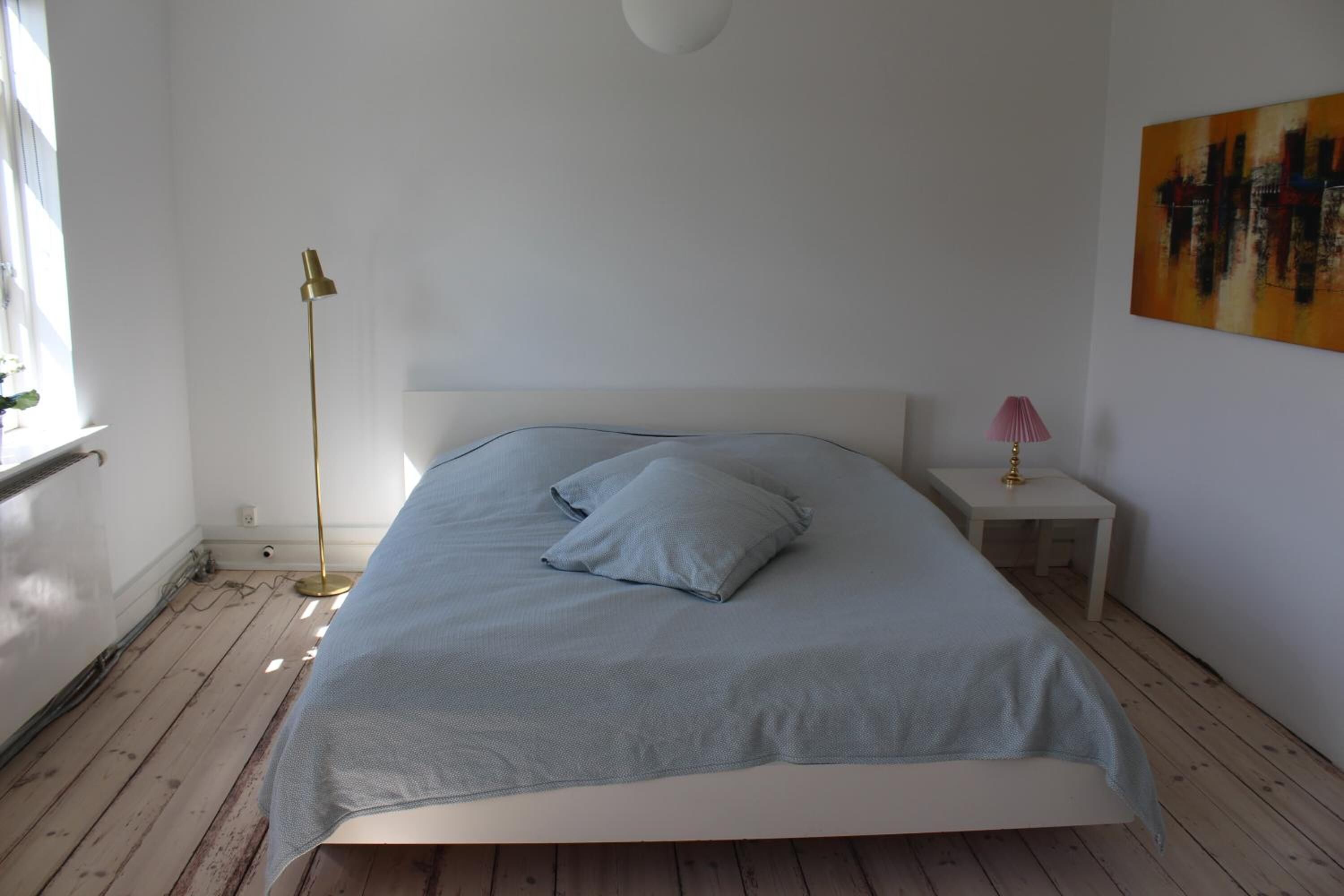 A bedroom with a bed, a nightstand, a floor lamp, and a painting on the wall.