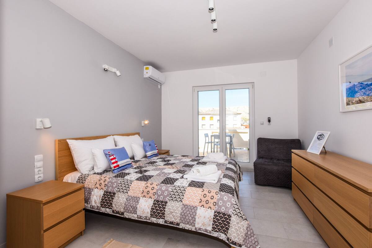 A bedroom with a bed, bedside tables, a dresser, a window, and an air conditioning unit.