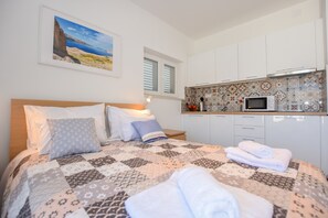 A bedroom with a bed, pillows, a quilt, a nightstand, a window, and a kitchen area with cabinets and a microwave. - Apartments Villa Magena (Pag)