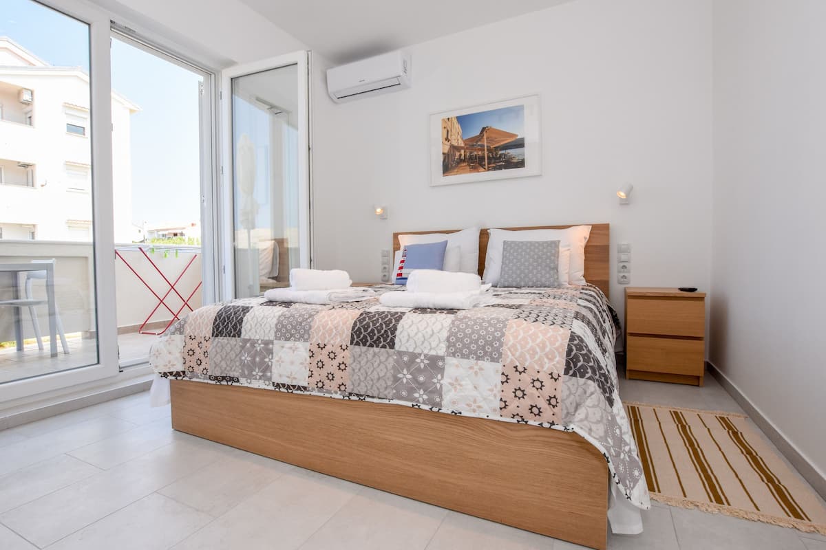 A modern bedroom with a large bed, a wooden nightstand, a small table, and a view through a sliding glass door.