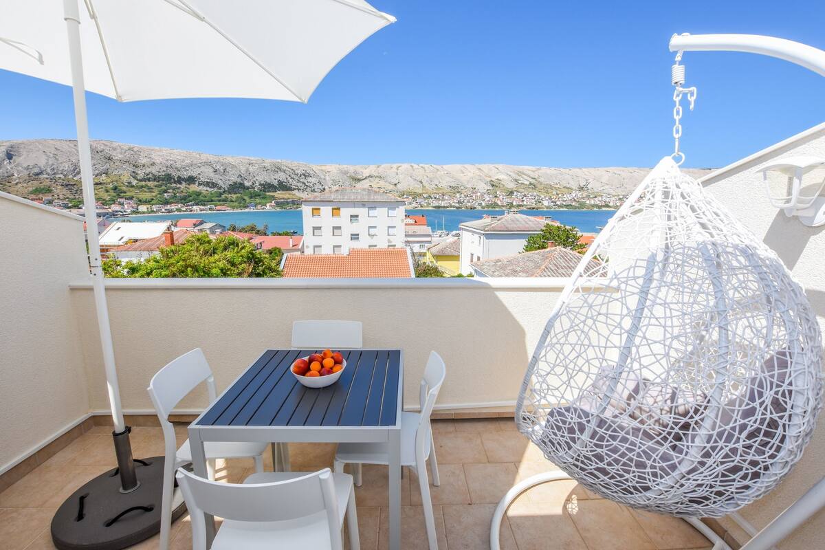 A balcony with a table, chairs, a hanging chair, and a view of a coastal town and sea.