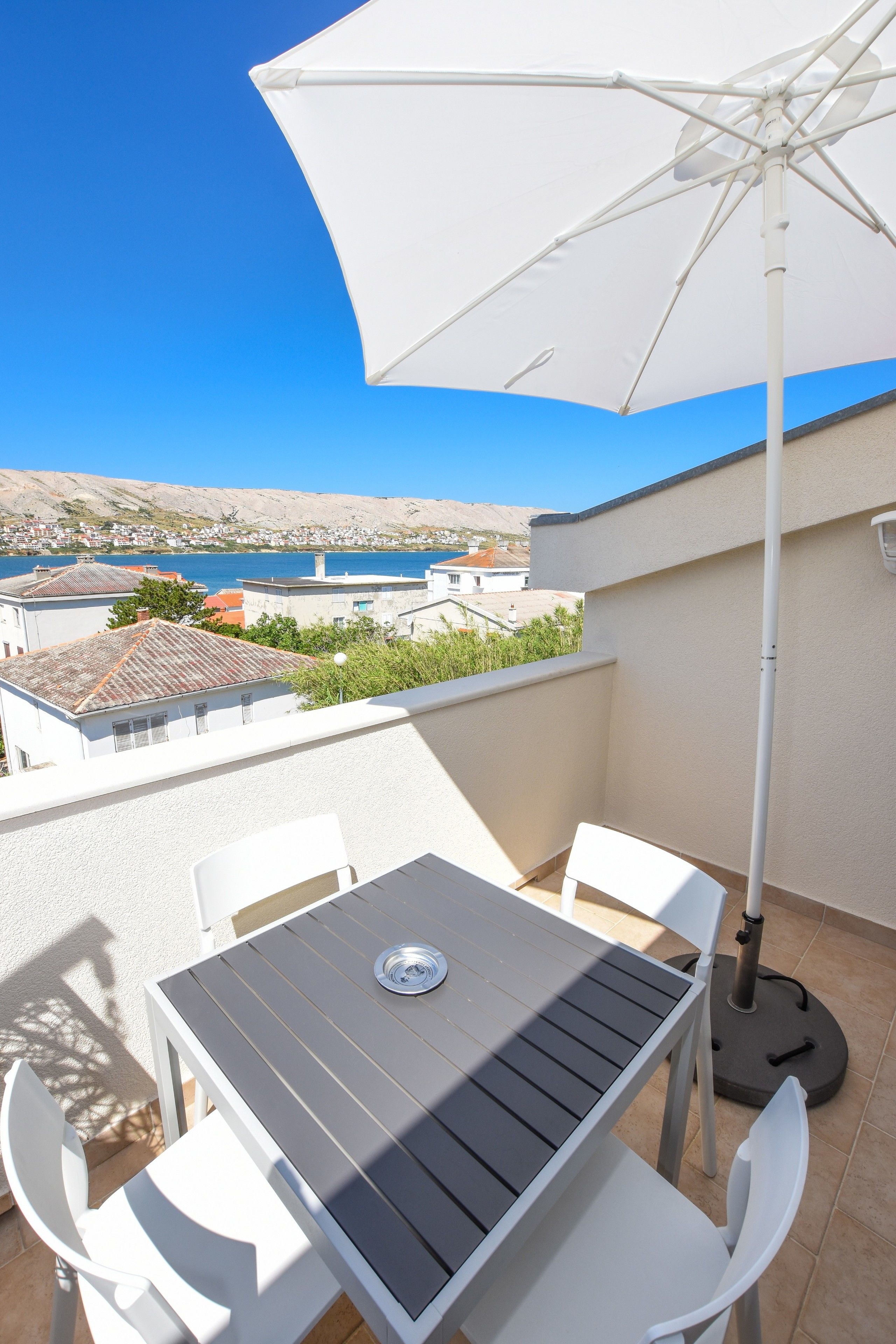A balcony with a table and chairs, an umbrella, and a view of a coastal town and sea.