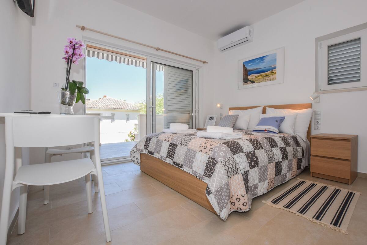 A bedroom with a bed, a desk, a chair, a bedside table, and a window with blinds.