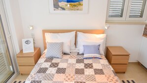 A neatly arranged bedroom with a large bed, two bedside tables, a wall-mounted air conditioner, a framed picture, and a window with shutters. - Apartments Villa Magena (Pag)