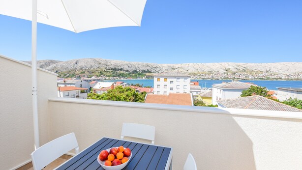 A balcony with a table and chairs, a bowl of fruit, and a view of a coastal town and mountains. - Apartments Villa Magena (Pag)