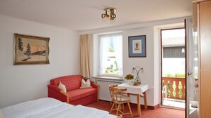 A room with a bed, a red sofa, a table with a chair, and a window with curtains. - Double room I - Guesthouse Zibert (Rottach-Egern)