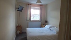 A bedroom with a bed, a bedside table, a lamp, a TV mounted on the wall, and a window with curtains.