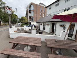 Un espacio al aire libre con mesas y bancos de madera, un edificio blanco con un paraguas rojo y un letrero que dice 'Watermans Arms'.