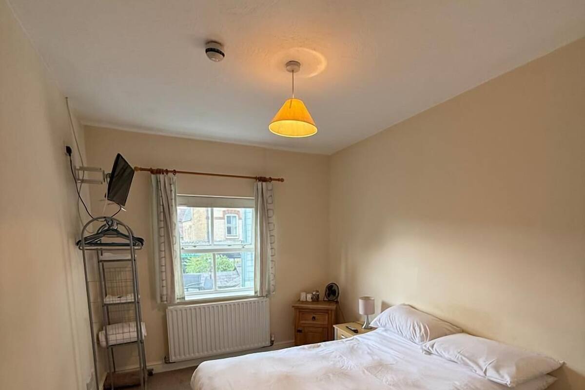 Een slaapkamer met een bed, nachtkastje, lamp, radiator, raam met gordijnen en een hanglamp.
