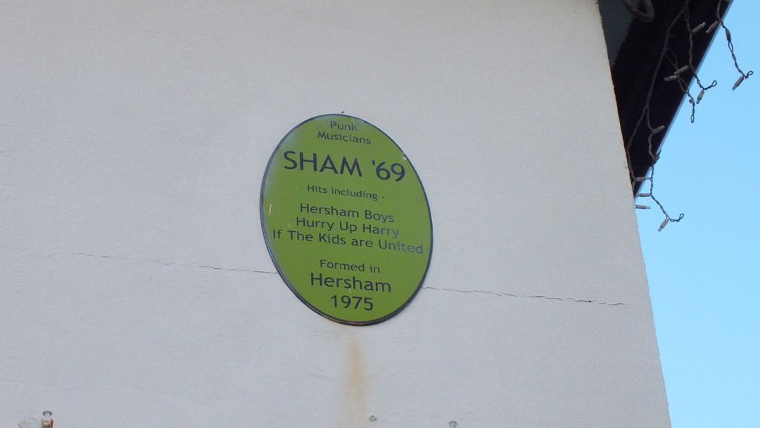 A green plaque on a building wall with text about a band named SHAM '69, formed in 1975 in Hersham.