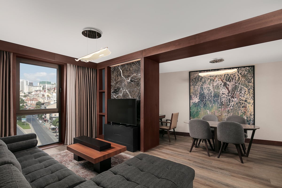 Una sala de estar moderna con un ventanal amplio, un televisor de pantalla plana, un comedor con mesa y sillas, y vistas a un paisaje urbano.