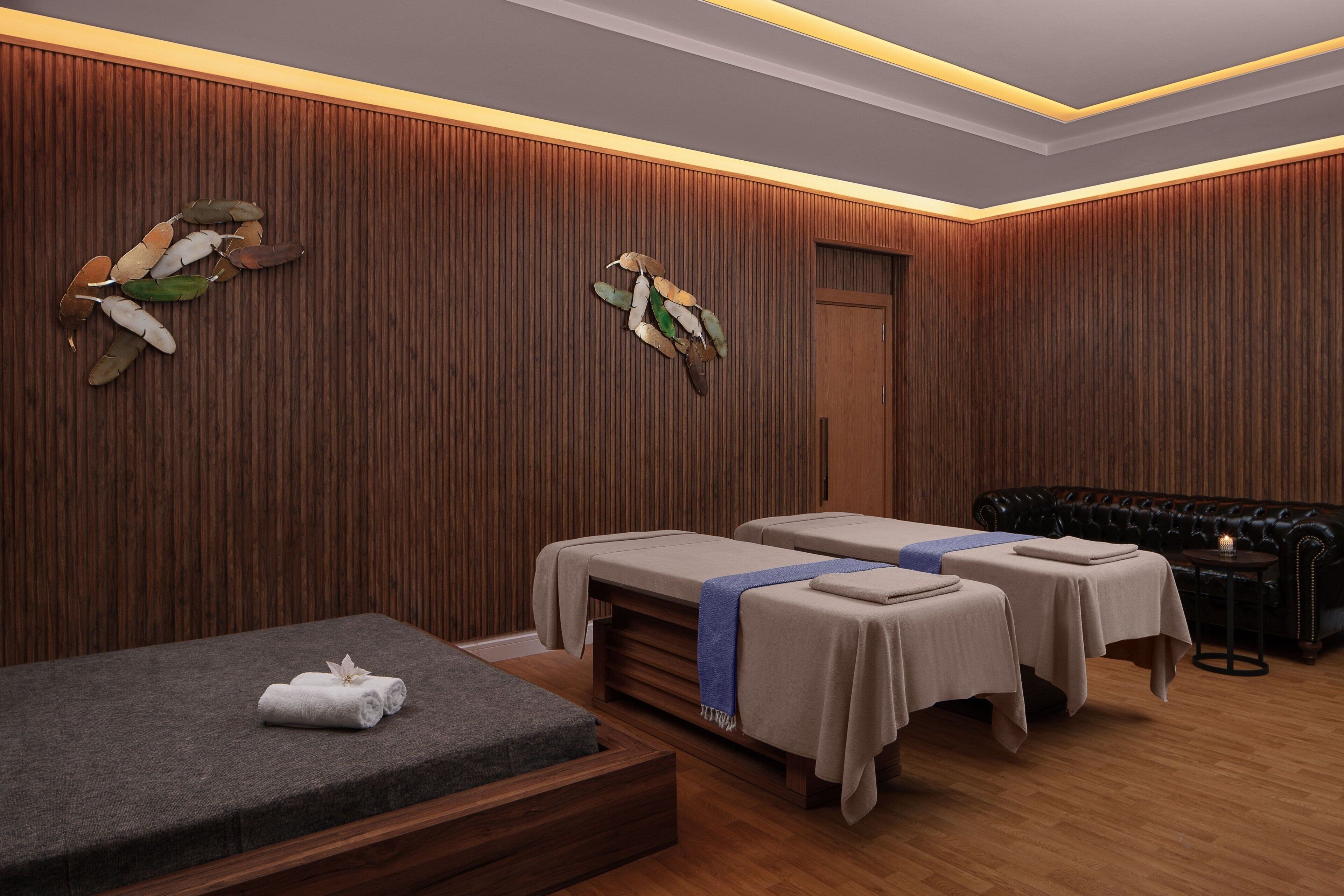A spa room with wooden paneling, two massage tables, a massage chair, and a small table with a candle.