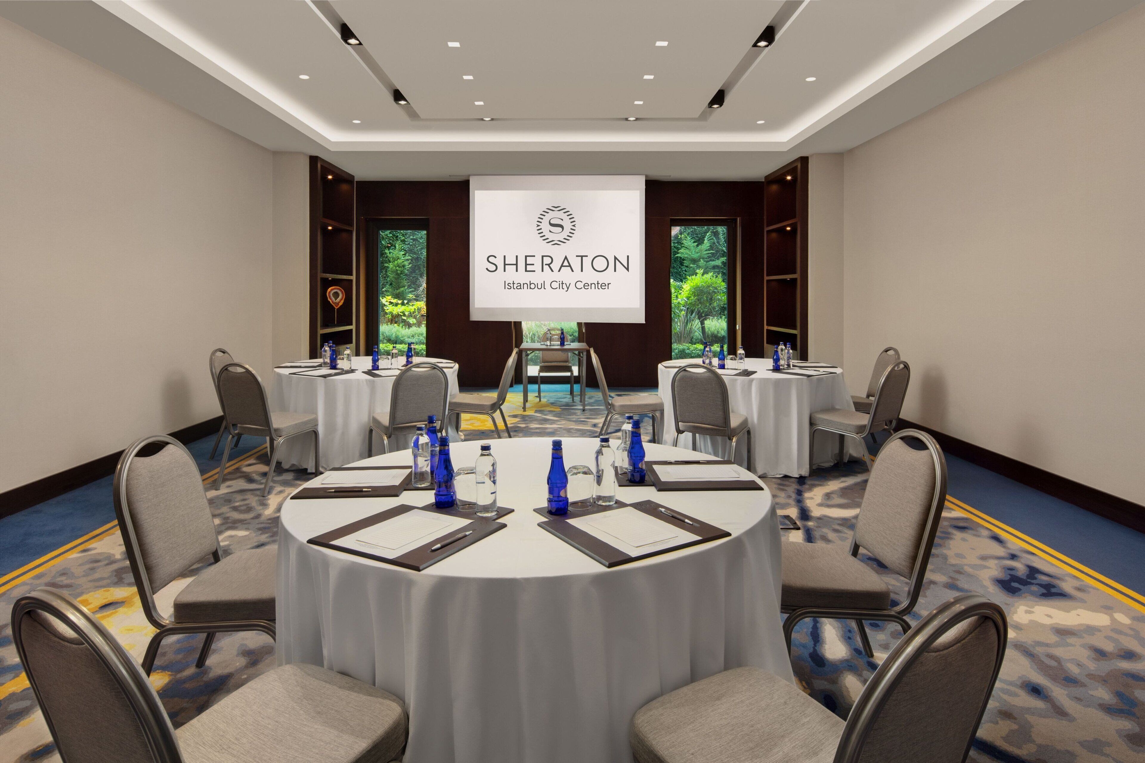 A Sheraton Istanbul City Center conference room with round tables, chairs, water bottles, and a large screen displaying the hotel's logo.