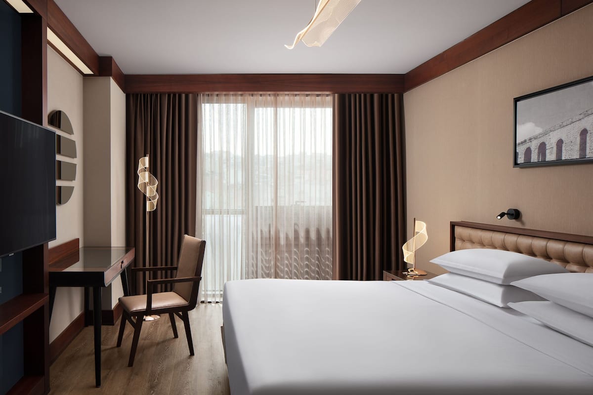 Habitación de hotel con cama, escritorio, silla, televisor y ventana con cortinas.