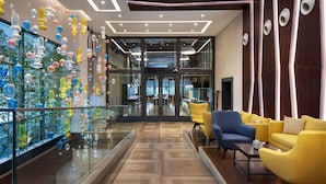 A modern lobby with a glass wall displaying colorful glass art, a seating area with yellow and blue chairs, and a wooden floor. - Sheraton Istanbul City Center (Istanbul)