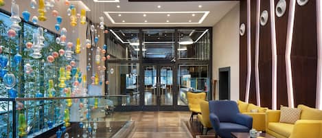 A modern lobby with a glass wall displaying colorful glass art, a seating area with yellow and blue chairs, and a wooden floor.