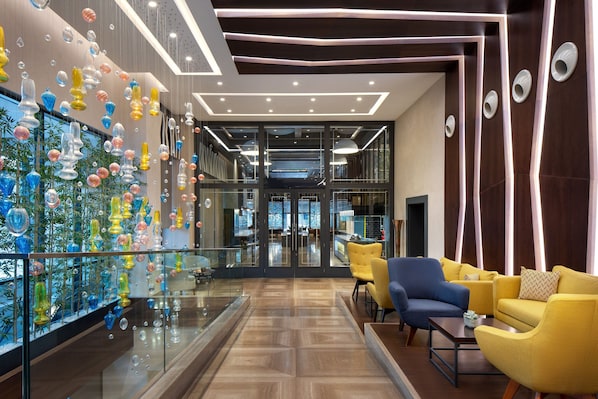 A modern lobby with a glass wall displaying colorful glass art, a seating area with yellow and blue chairs, and a wooden floor. - Sheraton Istanbul City Center (Istanbul)