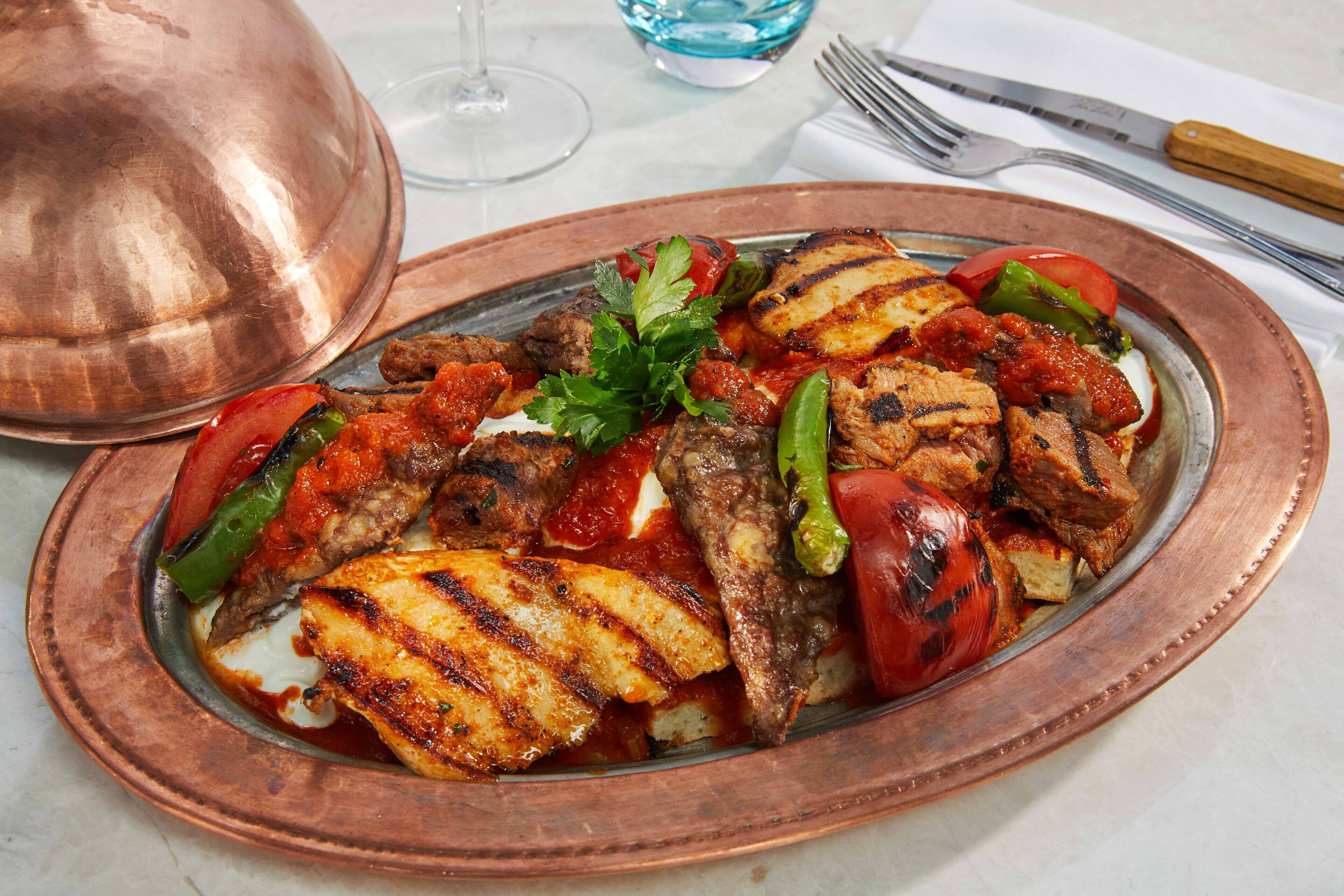 Un recipiente de cobre con carne a la parrilla, verduras y una guarnición sobre una mesa.
