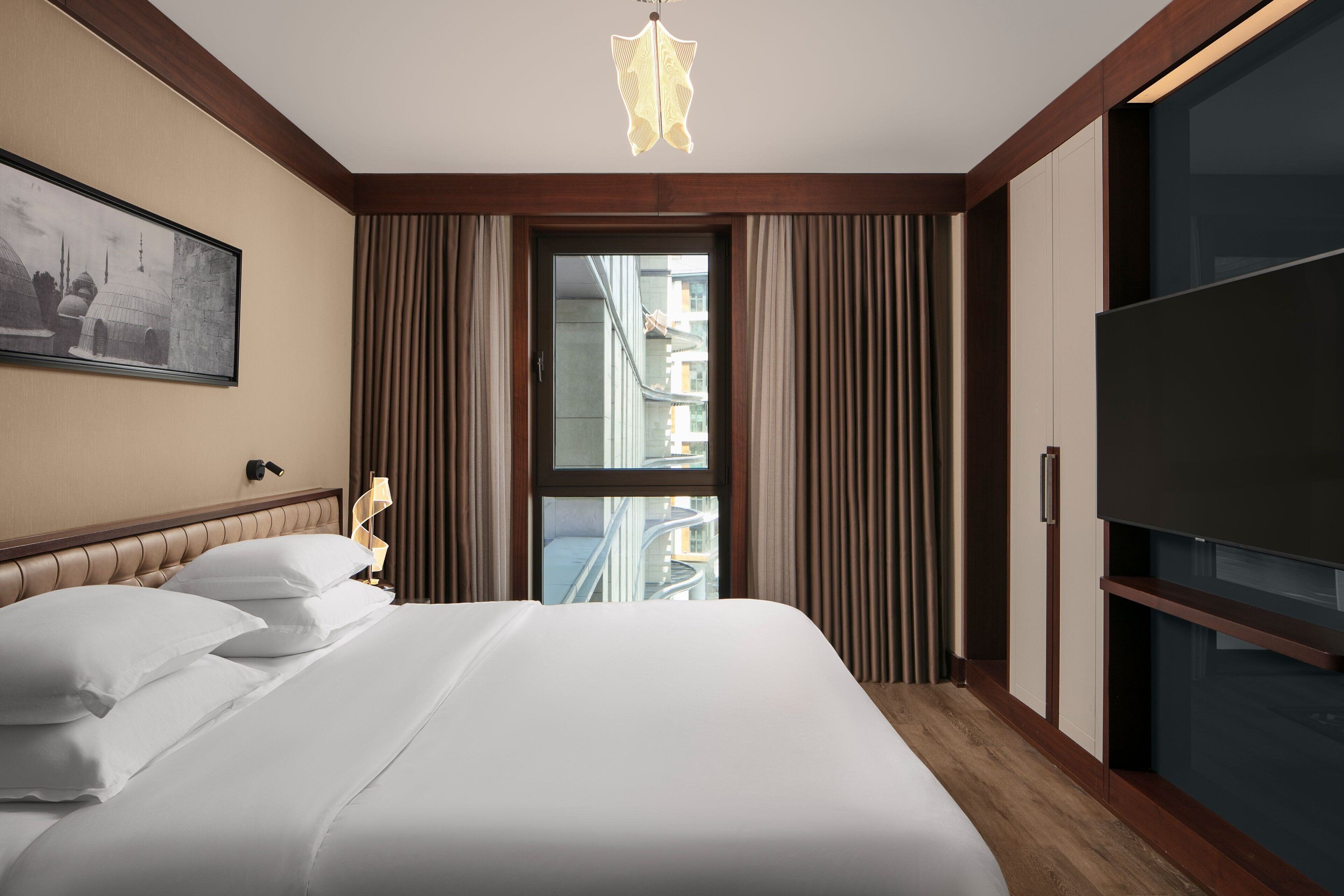 Quarto de hotel com uma cama grande, televisão, janela com cortinas e um quadro na parede.